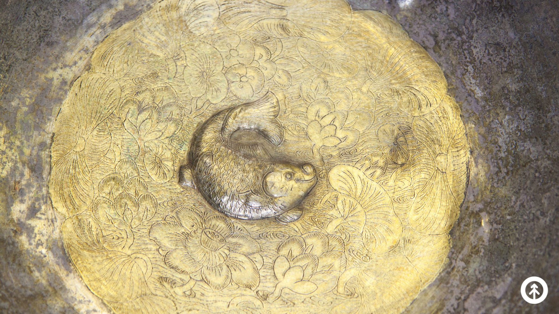 A circular bronze carving of a koi with lily pads and flowers. 