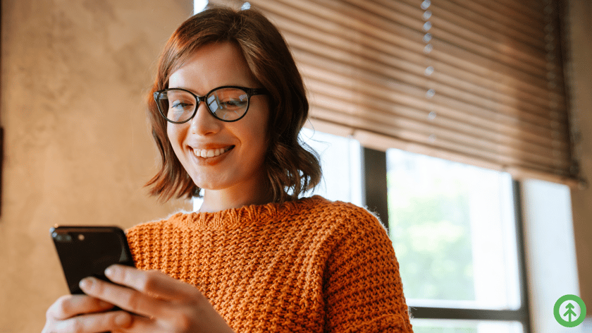 A person wearing glasses and an orange sweater smiles while viewing something positive on their mobile phone screen. 