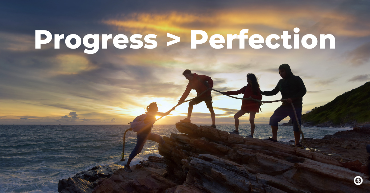 A group of people helping one person up a small rocky cliff by the ocean with a rope with text that reads “Progress > Perfection” from Growth Marketing Firm. 