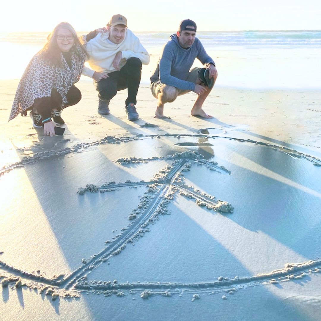Three Growth team members in front of a Growth logo drawn in the sand on a beach in Monterey, CA during a team retreat. 